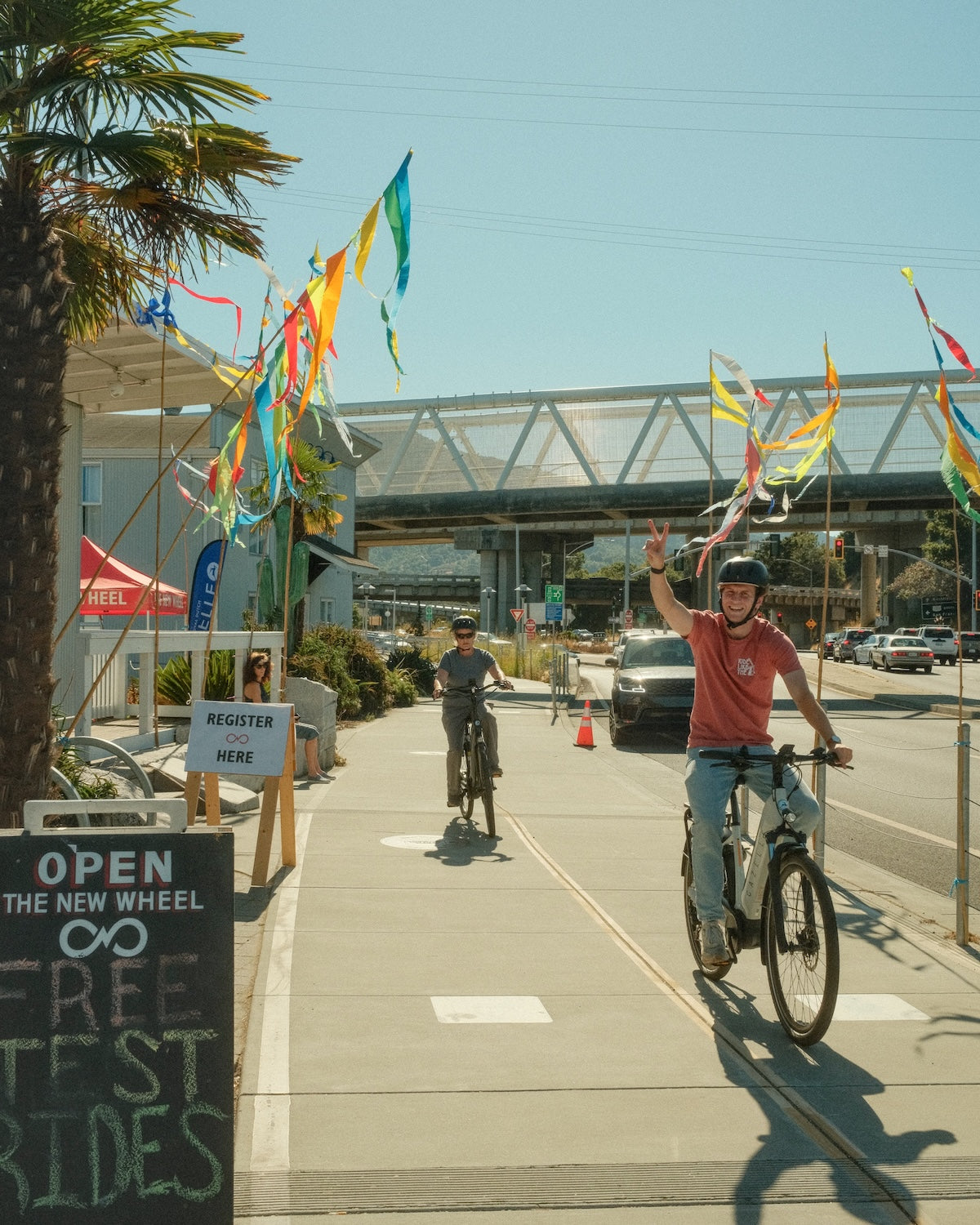 Two people riding bicycles on a sidewalk with colorful flags and a sign for free test rides.